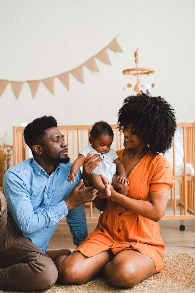 A imagem revela um casal negro sentado no chão com um bebê negro no colo, ambos olham afetuosamente para a criança. À esquerda o homem de  com barba e cabelos escuros, usa camisa azul clara e calça marrom, ao centro o bebe, usando body branco, a mãe tem cabelo cacheado black power na altura do ombro e usa vestido laranja. Fim da descrição