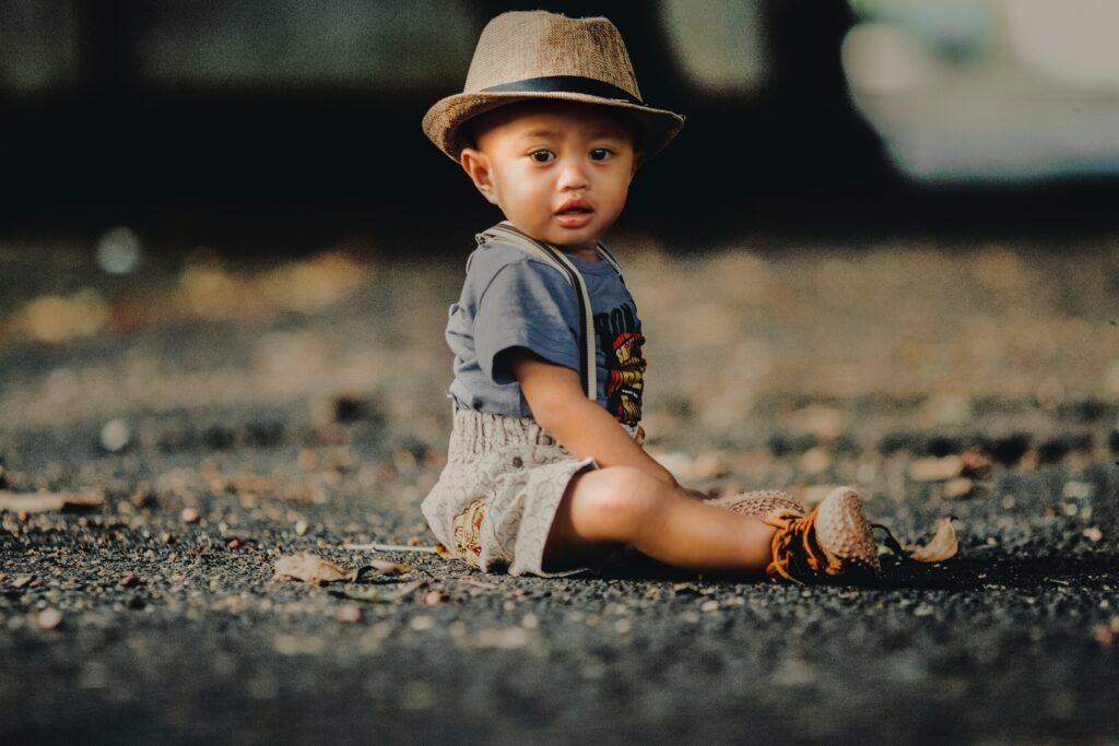 A imagem em ambiente externo com fundo desfocado, revela ao centro um bebe negro sentado no chão usando camiseta azul, shorts marrom claro do mesmo tom de seu chapéu estilo pananá. 