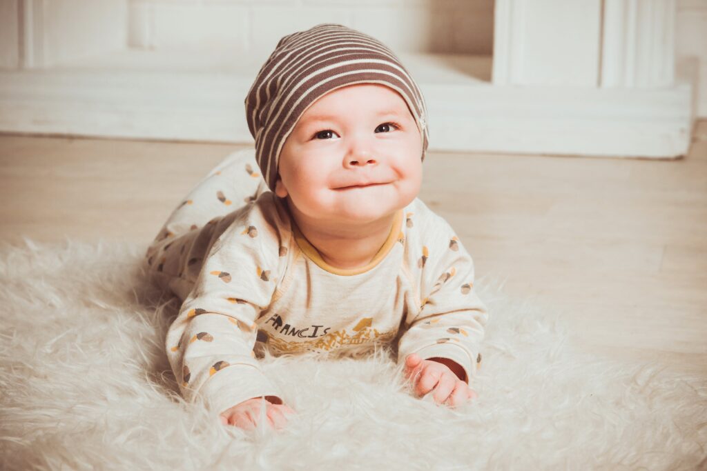 A imagem revela um bebe de pele clara deitado de barriga para baixo em um tapete branco feupudo. O bebe está com a cabeça inclinada para frente e se apoia com os antepraços no chão, usa uma touca marrom e macação bege com padronagem em marrom. Fim da descrição