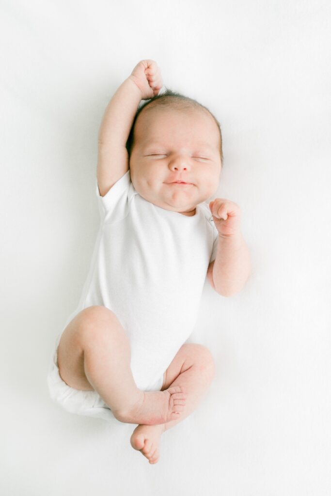 A imagem revela Bebê de bele clara deitado sob superficie branca com olhos fechados está usando um body branco, está com o braço direito acima da cabeça e esquedo dobrado ao lado do rosto, as pernas estão dobradas. Fim da descrição