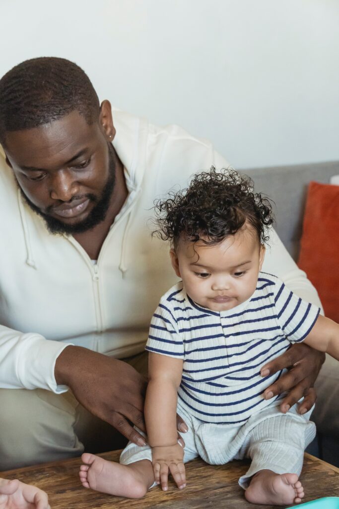 A imagem revela um homem negro com barba e cabelo curto, usando blusa cor creme e calça cinza clara, sentado no sofa com um bebe de aproximadamente 4 meses, o bebe está sentado em uma mesa de maneira, o homem apoia a criança segurando na cintura, o bebe usa camiseta listrada e calça cor creme. Fim dadescrição. 